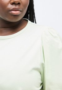 Κλείσιμο πλάνου ενός ατόμου με σκούρο δέρμα και κοτσίδες που φοράει γαλάζιο τοπ με φουσκωτά μανίκια, σε λευκό φόντο.