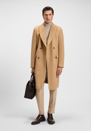 Abrigo de lana beige con solapas de muesca, cierre de doble botonadura; combinado con un jersey de cuello alto acanalado, pantalones ligeros beige y zapatos marrones.