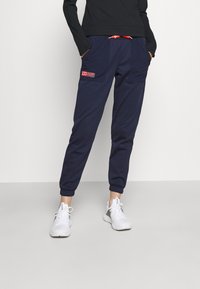 Navyblaue Jogginghosen mit elastischen Bündchen, zwei Seitentaschen und einem roten Kordelzug. Kombiniert mit weißen Sportschuhen und einem schwarzen Langarmoberteil.