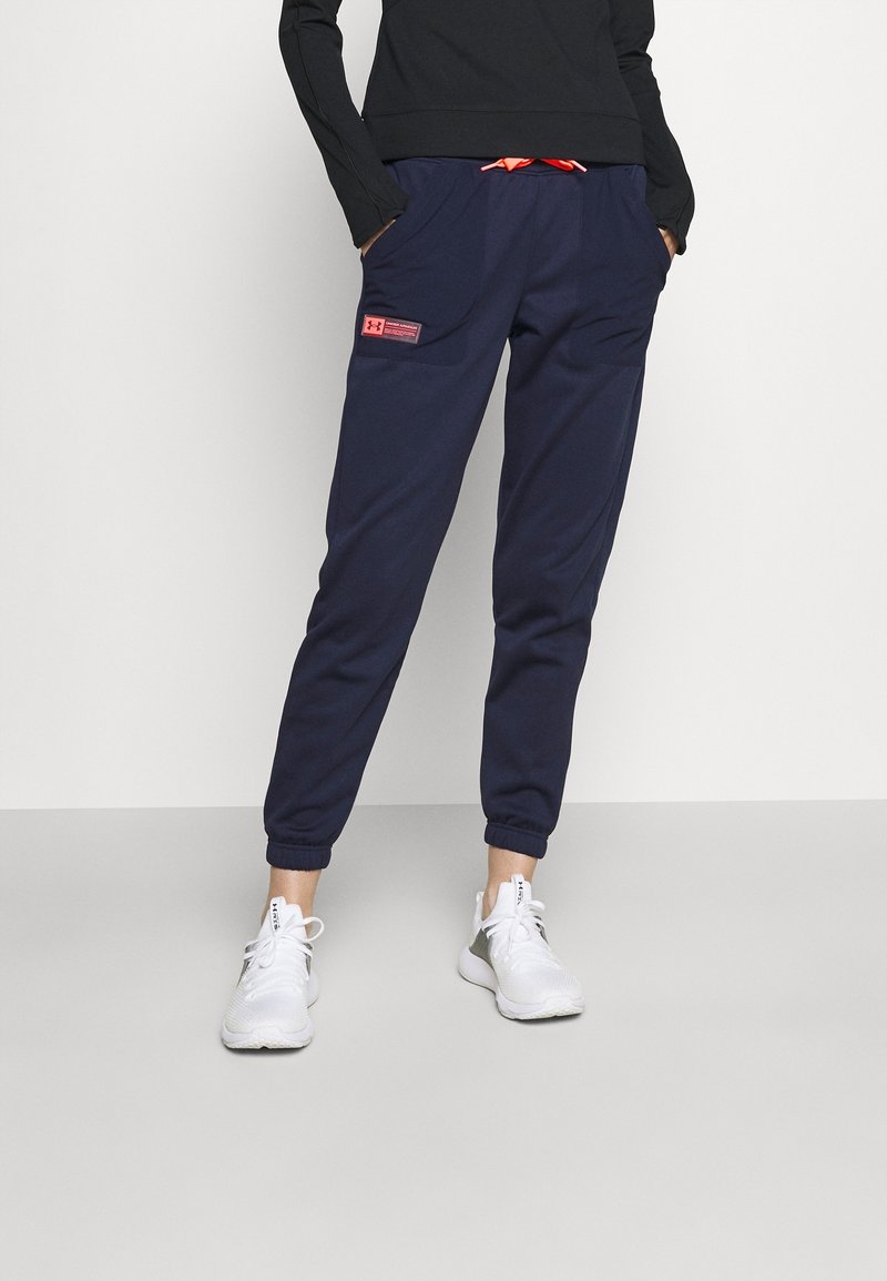 Navyblaue Jogginghosen mit elastischen Bündchen, zwei Seitentaschen und einem roten Kordelzug. Kombiniert mit weißen Sportschuhen und einem schwarzen Langarmoberteil.