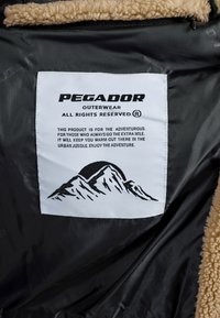 Interiorul moale din fleece maro cu o căptușeală neagră. O etichetă albă prezintă text negru și un grafic de munte, indicând informații despre marcă și produs.