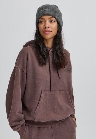 Liela brūna veste ar priekšējo kabatu, kapuci ar korķa auklām un melnu logotipu uz krūtīm. Valkāta kopā ar teksturētu pelēku adītu cepuri.