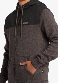 Brown zip-up hoodie with a dark gray upper panel. Features a drawstring hood, front pockets, and a logo patch on the chest.