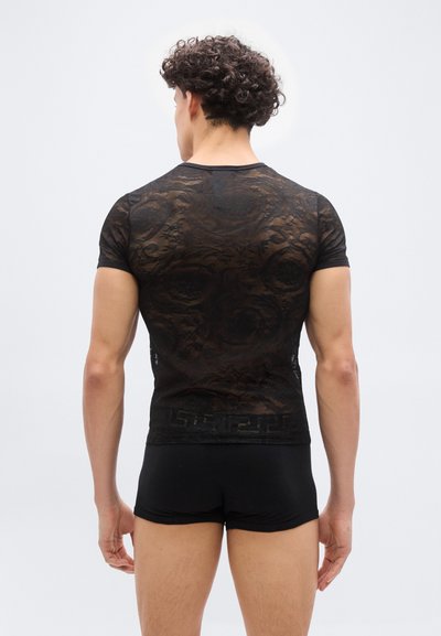 Άνδρας μοντέλο με σγουρά μαλλιά, φοράει μαύρο διαφανές πουκάμισο με μοτίβο λουλουδιών και κοντά μαύρα σορτς, γυρισμένος με την πλάτη σε λευκό φόντο.