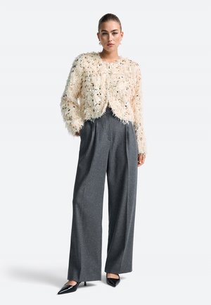 Cremfarbener, flauschig strukturierter, cropped Cardigan mit Pailletten, kombiniert mit grauen, weiten Hosen und spitzen, schwarzen Absätzen.