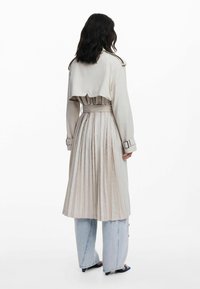 Light beige trench coat with pleated design, belted waist, and cuffed sleeves. Worn over light denim wide-leg pants.