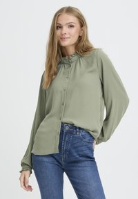 Long-sleeve, button-up blouse in olive green, featuring a ruffled collar and cuffs, paired with blue high-waisted jeans. Smooth fabric texture.