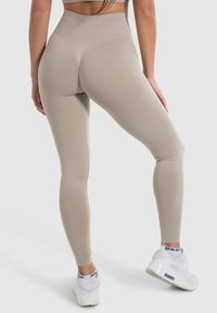 Frau in beigen, nahtlosen, hoch taillierten Leggings und weißen Sportschuhen, die mit leicht gespreizten Beinen vor einem einfarbigen Hintergrund steht.