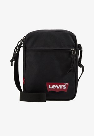 Čierna látková crossbody taška s nastaviteľným popruhom, predná priehradka na zips a červený detail loga Levi's na spodnej strane. Vysoký obdĺžnikový tvar.