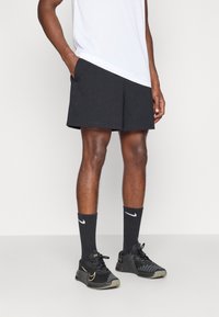 Μαύρα αθλητικά σορτς με πλάγιες τσέπες, συνδυασμένα με μαύρες κάλτσες και μαύρα αθλητικά παπούτσια με χρυσές λεπτομέρειες, φορεμένα με ένα λευκό πουκάμισο.