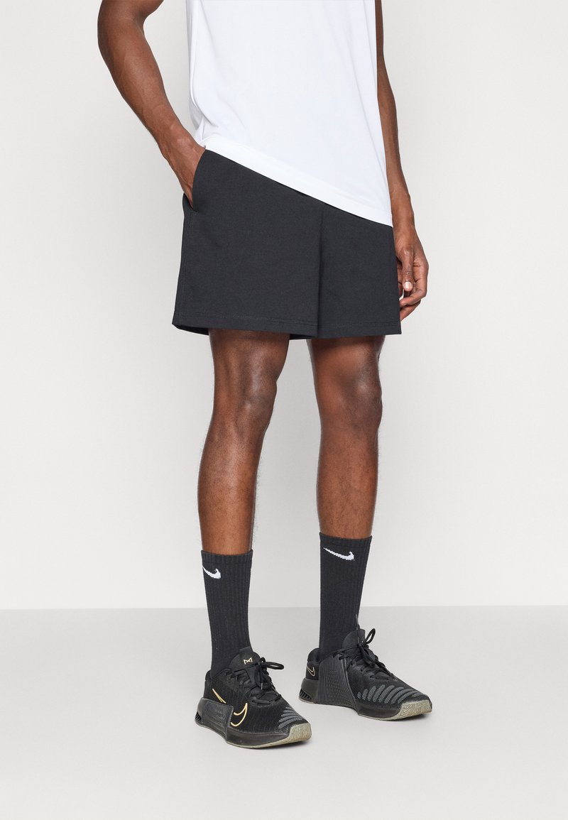 Μαύρα αθλητικά σορτς με πλάγιες τσέπες, συνδυασμένα με μαύρες κάλτσες και μαύρα αθλητικά παπούτσια με χρυσές λεπτομέρειες, φορεμένα με ένα λευκό πουκάμισο.