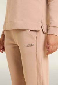 Sudadera de algodón color arena con un dobladillo en forma de alta-baja y aberturas laterales, combinada con pantalones de chándal a juego que incluyen un bolsillo y la palabra 'FREDDY' en strass.