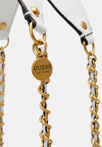 Hvid faux læderrem med sorte accenter, guld metalhardware, snoet kæde detaljer og et rundt mærke med indskriften "GUESS EST.1981."