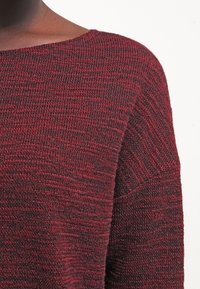 Primo piano di una persona che indossa un maglione a maglia tessuta di colore bordeaux e nero con un ampio scollo a barca.