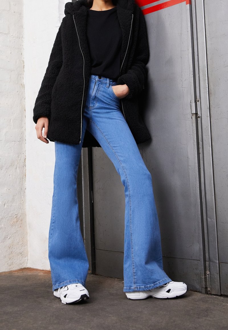 Giacca a zip foderata in pile nera, jeans blu a zampa d'elefante e sneaker bianche. La giacca ha un cappuccio e una vestibilità ampia, mentre i jeans sono a vita alta.