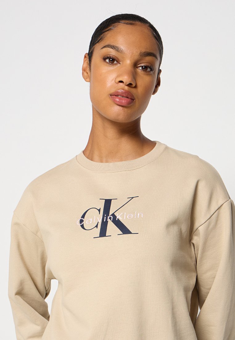 Beżowy długi rękaw sweatshirt o prążkowanej teksturze, z wyraźnym granatowym logo CK i białym napisem "Calvin Klein" na klatce piersiowej.