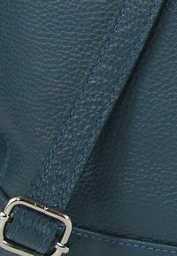 Detail van een donkerblauwe leren tas met een gestructureerd oppervlak, contrasterende blauwe stiksels en een glanzende zilveren gesp op de band.