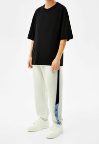 Osoba oblečená v volném černém tričku, bílých teplácích s černými a modrými bočními pruhy a bílých teniskách, stojící na jednobarevném pozadí.