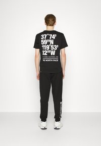 Svart kortärmad t-shirt med stora vita bokstäver som visar koordinater och "THE NORTH FACE"-logga; kombinerad med svarta sweatpants och sneakers.