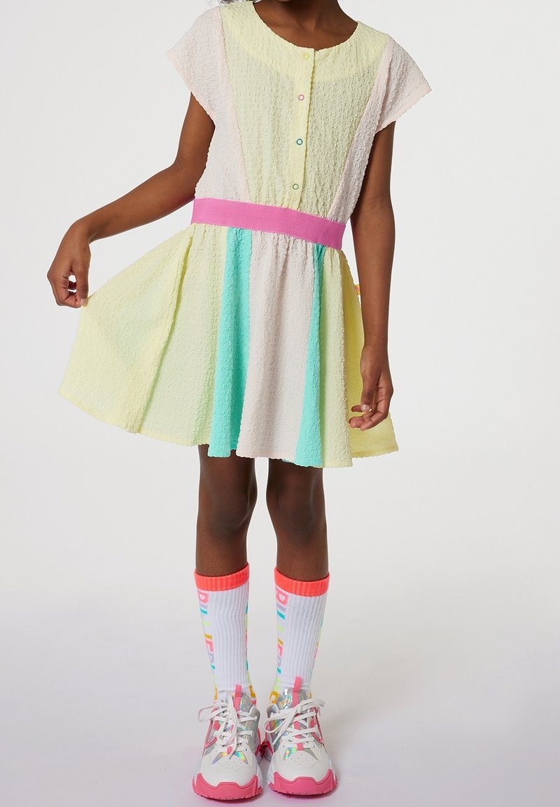 Enfant portant une robe jaune pastel, rose et turquoise avec une ceinture rose, des chaussettes hautes colorées et des baskets blanches avec des semelles roses.