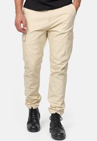Pantaloni cargo beige con multiple tasche laterali, vestibilità affusolata, realizzati in tessuto leggero. Indossati con stivali neri stringati.