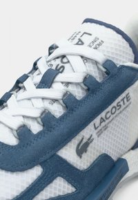 Λευκά αθλητικά παπούτσια από πλέγμα με μπλε σουέντ λεπτομέρειες, έντονη κορδόνια, σήμανση και υφή στη σόλα. Περιλαμβάνεται χαρακτηριστική λεπτομέρεια λογοτύπου.