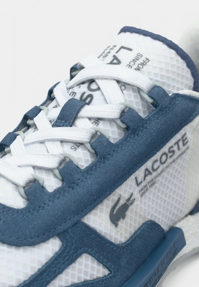 Baskets en maille blanche avec des accents en daim bleu, présentant un laçage prononcé, un logo bien visible et une semelle texturée. Détail du logo notable inclus.