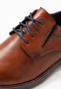 Zapato de cuero marrón con cordones azul marino, con un acabado suave, detalles de costura sutiles y suela de goma. Diseño clásico de cordones.