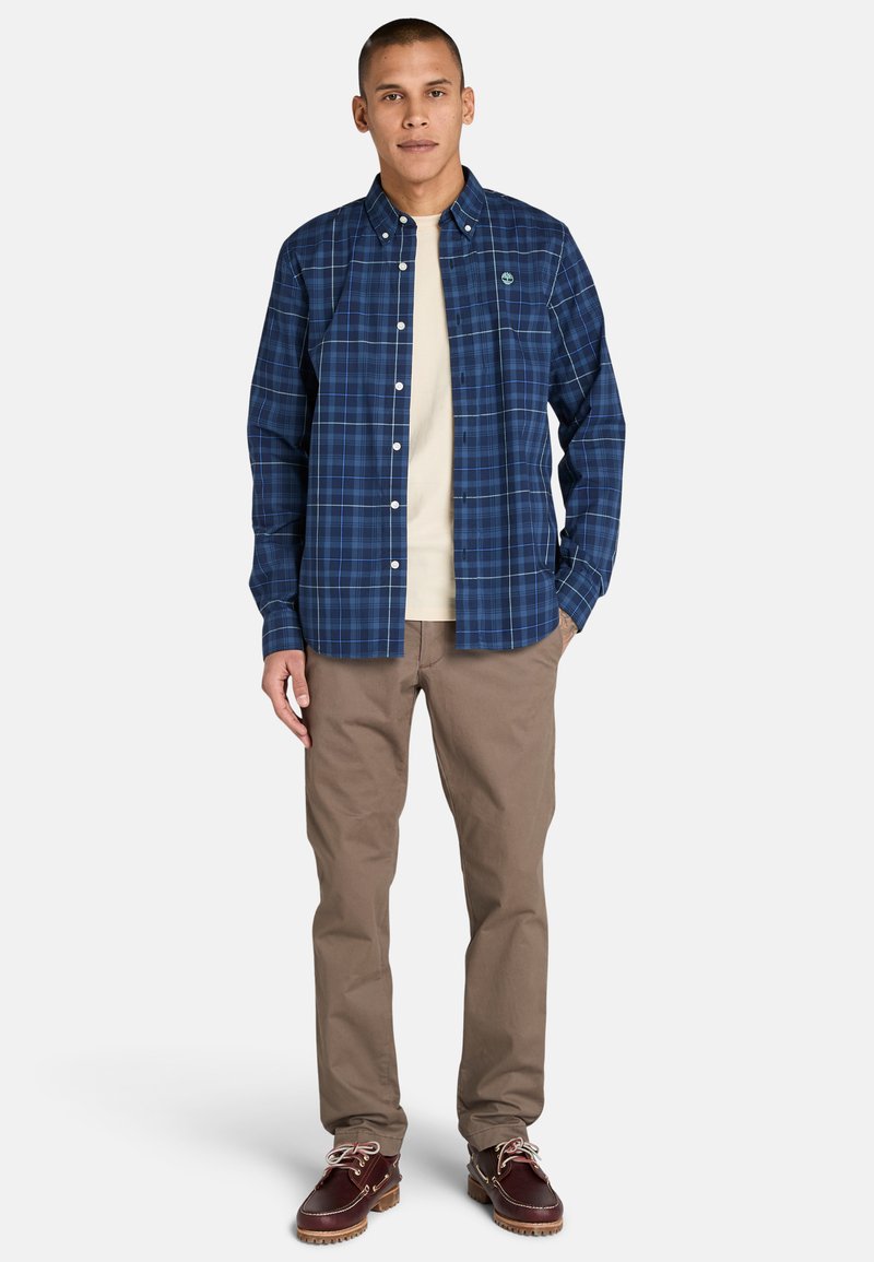 Blaues kariertes Button-Down-Hemd mit leichtem Glanz, kombiniert mit einem cremefarbenen T-Shirt, beigen Hosen und dunkelbraunen Bootsschuhen mit Schnürsenkeln.