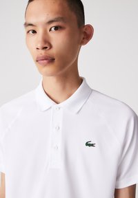 Vit polo-skjorta med en grön broderad krokodil-logotyp på vänster bröst, tillverkad av texturerad bomullsög och med klassisk krage.