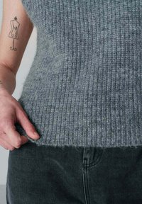 Grijze, ribbelige gebreide trui met een zachte textuur, gedragen over donkere denim. De zoom van de trui ligt net boven de taille en benadrukt het aansluitende ontwerp.