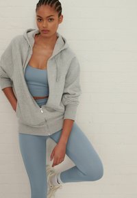 Lichtgrijze hoodie met ritssluiting en een relaxed fit, gecombineerd met een blauwe sport-bh en bijpassende blauwe leggings. Model draagt witte sneakers.