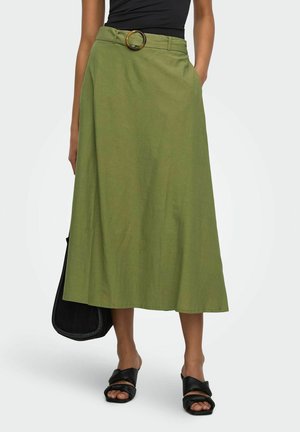 Woman wearing a green midi skirt with a large round belt buckle, black slide sandals, holding a black bag, standing on white background.