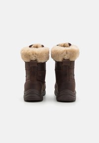 Een paar donkerbruin leren winterlaarzen met dikke beige bontvoering, gezien vanuit de achterkant op een witte achtergrond.