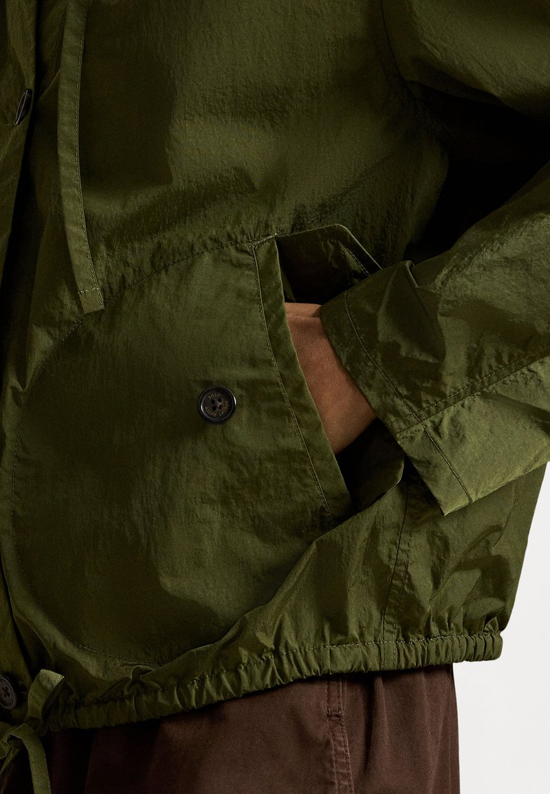 Hand tucked into large, curved pocket of olive green jacket with button detail and elastic hem, worn over brown pants.