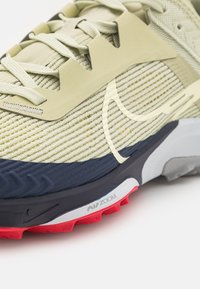 Gros plan d’une chaussure de course beige et bleu marine avec tige en mesh, logo blanc Nike, semelle rouge et logo Air Zoom sur la semelle intermédiaire.