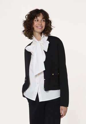 Femme aux cheveux bouclés souriante, portant un chemisier blanc avec un grand nœud, une veste noire et un pantalon, sur un fond uni.