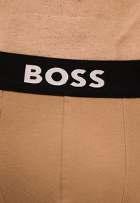 Ceinture de sous-vêtements beige avec bande élastique noire portant le texte blanc "BOSS", portée sur peau nue.