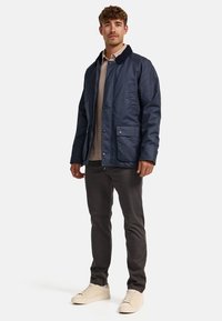 Veste bleu marine avec fermeture à bouton, deux poches avant et un col doux. Portée sur une chemise beige et associée à un pantalon sombre et des baskets claires.