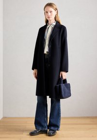 Femme portant un long manteau noir sur une chemise rayée et un jean évasé, tenant un sac à main texturé bleu marine, se tenant sur un sol en bois.