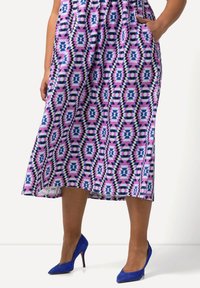 Lila gemustertes Midikleid mit geometrischen Formen, ausgestattet mit Taschen und einem fließenden Design, kombiniert mit blauen High Heels.
