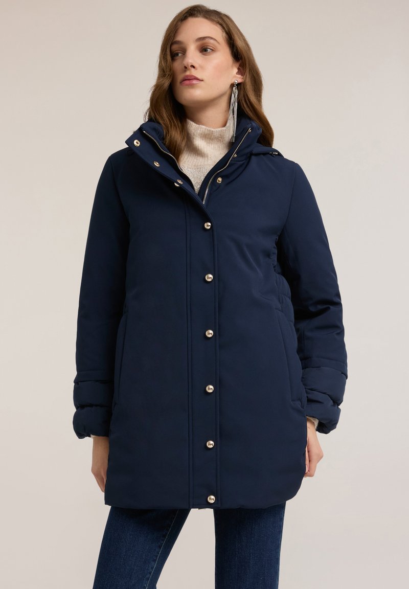 Abrigo acolchado azul marino con un diseño entallado, que presenta un cuello alto, botones de metal dorado y bolsillos laterales. La textura parece suave e aislante.