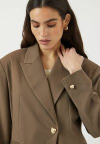 Brun oversized blazer med notchad krage, två guldhjärtformade knappar och en strukturerad tyg. Modellen rör vid sin nacke i en subtil pose.