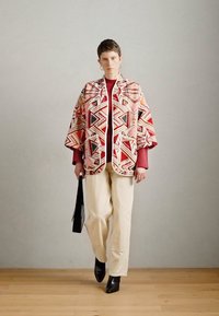 Többszínű mintás köpeny geometriai mintákkal, piros felső és krémszínű nadrág felett. Fekete magas sarkú csizmák és egy fekete táska egészíti ki a megjelenést.