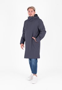 Cappotto grigio con cappuccio, maniche lunghe, chiusura frontale con zip e tasche laterali. Indossato sopra jeans blu e sneaker bianche. Tessuto liscio e opaco.