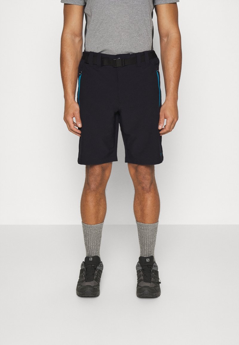 Homme portant un short noir avec des fermetures éclair bleues, des chaussettes grises, des chaussures de randonnée noires et un t-shirt gris, debout devant un fond blanc.
