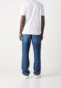 Wit katoenen T-shirt en loszittende blauwe spijkerbroek met achterzakken en subtiele stikseldetails, gecombineerd met lichte schoenen.