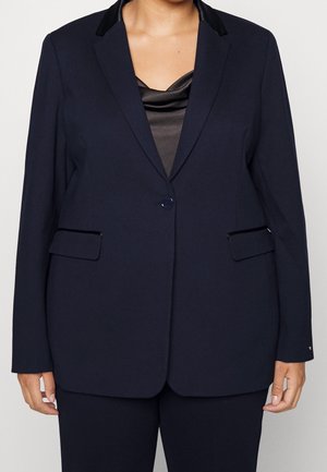 Personne portant un blazer noir ajusté avec des revers en satin et un pantalon assorti par-dessus un chemisier en satin noir, debout devant un fond uni.