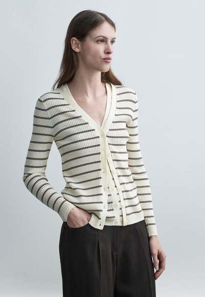 Jeune femme portant un cardigan crème avec de fines rayures horizontales noires et un pantalon noir, debout avec une main dans la poche, regardant vers la droite.