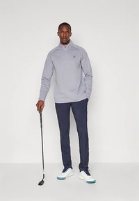 Hombre con pulóver gris de media cremallera, pantalones azul marino y zapatos de golf blancos, de pie con un palo de golf frente a un fondo liso.